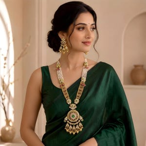May include: A long gold-toned necklace featuring multiple strands of white beads, intricate metalwork, and a large circular pendant with green and red accents. Matching gold-toned drop earrings with similar bead and stone detailing are worn.