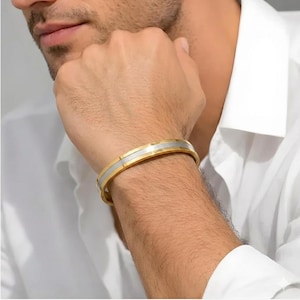Puede incluir: Un brazalete dorado y plateado. El brazalete tiene una banda dorada con una franja central plateada. El brazalete se lleva en la muñeca.