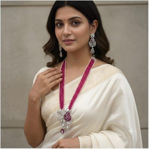 May include: A long necklace featuring multiple strands of small, vibrant pink beads accented with white pearls. The pendant is a large, silver-toned floral design encrusted with clear stones and a central pink stone. Matching drop earrings are included.