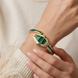 May include: A gold bracelet in a snake design, adorned with emerald green and clear gemstones. The bracelet wraps around the wrist, with a detailed head and tail. The gemstones are set in a repeating pattern.