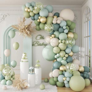 May include: A baby shower display with a balloon arch in green, gold, and white. The words "Oh Baby" are visible. The scene includes a cake, pedestals, and balloons, creating a celebratory atmosphere. The balloons are in various sizes.