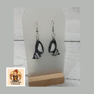 May include: A pair of butterfly wing earrings. Each earring features a black and white wing design with silver-colored hooks. The earrings are displayed on a white stand with a wooden base. The logo "Clay'd and Confused" is visible in the lower left corner.
