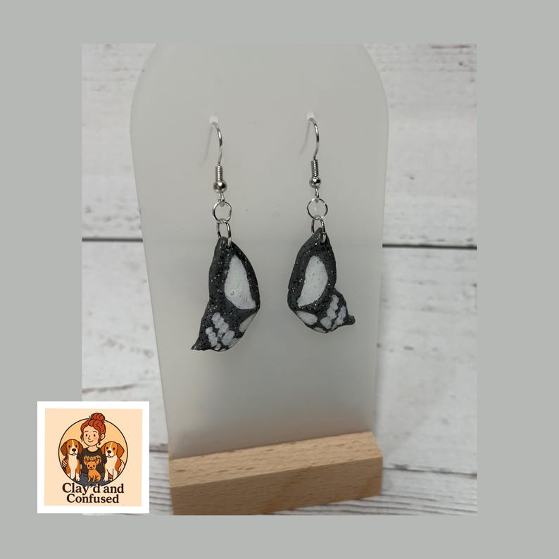 May include: A pair of butterfly wing earrings. Each earring features a black and white wing design with silver-colored hooks. The earrings are displayed on a white stand with a wooden base. The logo "Clay'd and Confused" is visible in the lower left corner.