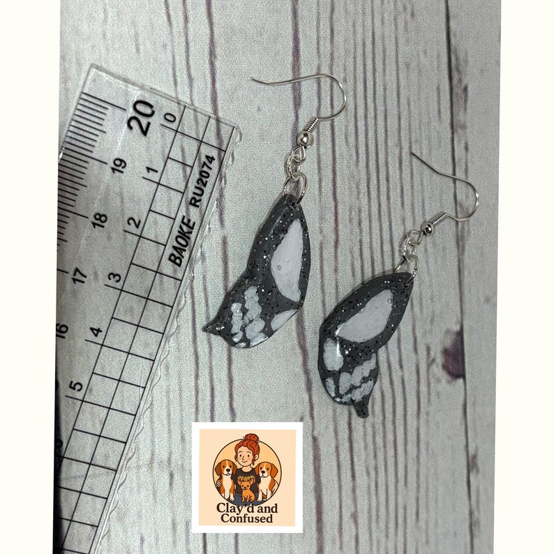 May include: A pair of handmade butterfly wing earrings. The wings are black with white accents and silver-colored ear wires. A ruler with metric measurements is in the background. The earrings are from Clay'd and Confused.