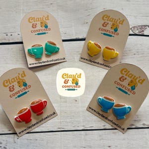 May include: Four pairs of coffee cup stud earrings in teal, yellow, red, and blue, each with a brown coffee fill. The earrings are presented on tan cards with the text "Clay'd & Confused" and a website address.