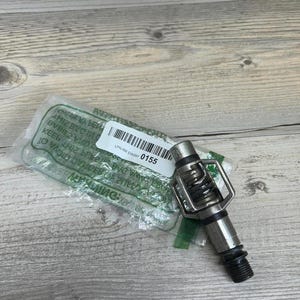 May include: A silver bicycle pedal with a black threaded end, a spring, and a metal cage. The pedal is partially visible inside a clear plastic package with a white label that reads "LPN RR EW287 0155". A yellow tape measure is in the background.