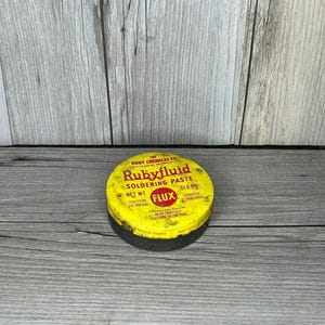 May include: A round, yellow and black Rubyfluid soldering paste tin. The lid features the text "Rubyfluid SOLDERING PASTE" and "FLUX". The net weight is 51.6 grams. The tin is on a wooden surface.