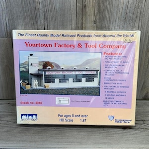 以下が含まれることがあります： 「Yourtown Factory & Tool Company」の模型鉄道キットボックス。ボックスは主に紫と白で、工場の画像があります。「6色成形」と「HOスケール1:87」の文字が含まれています。