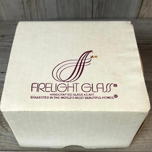 May include: A cream-colored box with the words "FIRELIGHT GLASS" in a burgundy font. The box also features a stylized burgundy "f" design and the text "HANDCRAFTED GLASS AS ART COLLECTED IN THE WORLD'S MOST BEAUTIFUL HOMES."