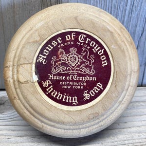 Puede incluir: Un recipiente redondo de madera clara para jabón de afeitar. El centro presenta una etiqueta burdeos con las palabras "House of Croydon" y "Shaving Soap" en una fuente decorativa, junto con un diseño de escudo. El distribuidor se indica como Nueva York.