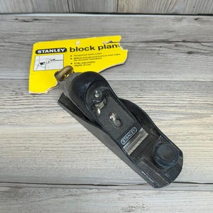 Puede incluir: Un cepillo de bloque Stanley negro con un botón de ajuste dorado y el logotipo de Stanley. La tarjeta de información del producto amarilla dice "block plan" y enumera las características. La herramienta está sobre una superficie de madera clara.