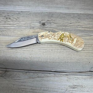 May include: A folding knife with a cream-colored handle featuring a detailed relief carving of a mountain scene and a gold-colored figure. The blade is silver and bears engraved text. The knife is open and ready for use.