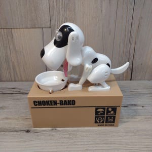 May include: A white and black dog-shaped coin bank with a pink tongue, a small white bowl, and the text "CHOKEN-BAKO" on the front. The bank is made of plastic and sits on a brown box. The bank is marked "MADE IN CHINA".