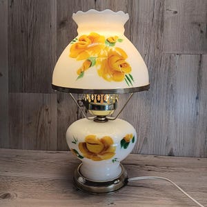 May include: A vintage white glass lamp with a floral design featuring yellow roses and green leaves. The lamp has a metal base and a white lampshade with a scalloped edge. The lamp is plugged in and lit.