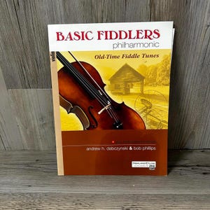 Puede incluir: Un libro titulado "BASIC FIDDLERS philharmonic" con "Old-Time Fiddle Tunes" en la portada. La portada presenta un violín y una imagen sepia de una escena rural. El libro es de Andrew H. Dabczynski & Bob Phillips.