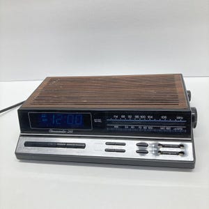 May include: A vintage Chronomatic-268 digital alarm clock radio with a brown wood-grain top and a black base. The clock displays the time in blue digits and has FM/AM radio settings. The clock measures approximately 8 inches wide.