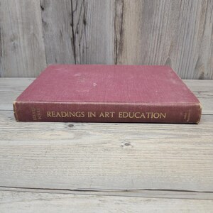 Puede incluir: Un libro antiguo titulado "Readings in Art Education" con una cubierta granate. El título del libro y el nombre del autor están impresos en letras doradas. El libro está sobre una superficie de madera clara.