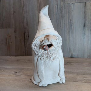 May include: A handcrafted, off-white ceramic gnome figurine with a long, pointed hat and a full, textured beard. The gnome has a detailed face and is holding a small, colorful object in its mouth. The figurine is standing on a wooden surface.