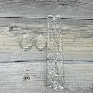 May include: Two clear, teardrop-shaped earrings and a rectangular, textured clear glass object. The earrings have a small loop at the top for hanging. The glass object has a textured surface and a rectangular shape.