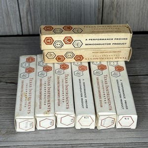 May include: Several vintage Texas Instruments semiconductor product boxes are stacked and arranged on a wooden surface. The boxes are primarily white and tan, with the company name and product information printed in black and orange. The boxes are labeled "A Performance Proved Semiconductor Product."