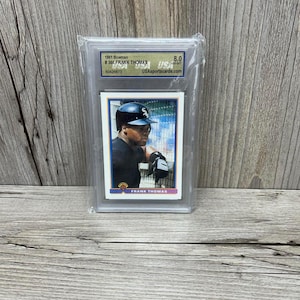 Könnte beinhalten: Eine 1991 Bowman Baseballkarte von Frank Thomas, bewertet mit 8,0 von USAsportscards.com, in einer Schutzhülle aus Kunststoff. Die Karte zeigt Thomas in seiner Chicago White Sox Uniform vor blauem Hintergrund mit weißem Rand.