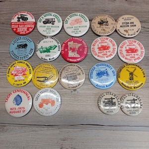 May include: A selection of vintage round pin badges with diverse designs and text. The badges feature illustrations of tractors, steam engines, and event names such as "Steam-Gas Museum Show" and "Pioneer Power Show." Colours include red, blue, yellow, and white.