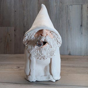 May include: A handcrafted ceramic garden gnome statue. The gnome is wearing a white pointed hat and a white robe. It has a long white beard and is holding a cigar in its mouth. The gnome's face is painted with pink and brown tones.