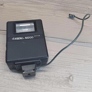 May include: A black Focal M200 camera flash with a rectangular form and a clear flash tube. The flash has a grey cord attached. The text "FOCAL M200" and "20-01-38" are printed on the front.