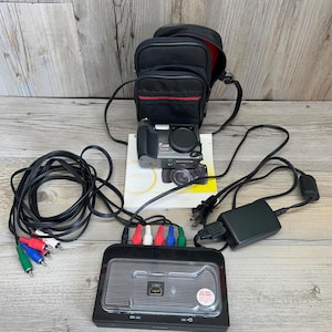 May include: A black digital camera with a black carrying case, a charger, and various cables. The camera is on top of a Kodak Z1012 IS manual. The carrying case has a red stripe.