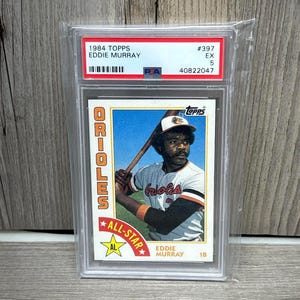 Puede incluir: Una tarjeta de béisbol Topps de 1984 de Eddie Murray en una funda protectora. La tarjeta muestra a Murray con su uniforme de los Orioles, sosteniendo un bate. La tarjeta está calificada como EX 5 e incluye el texto "ALL-STAR".