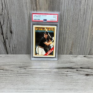 Puede incluir: Una tarjeta de béisbol de Sid Bream Topps Tiffany de 1988, calificada como Mint 9, en un soporte protector. La tarjeta muestra a un jugador de béisbol con el uniforme de los Piratas, con el nombre del equipo. Fondo de madera.