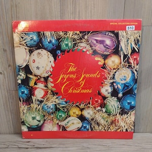 May include: Vintage vinyl record album cover titled "The Joyous Sounds of Christmas." The cover features a red border and a central design of Christmas ornaments in various colors, including blue, gold, and red. The text "Special Collectors Edition" is visible.