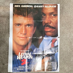 May include: A movie poster for "Lethal Weapon 2" featuring Mel Gibson and Danny Glover. The poster has a split design with the actors' faces on either side. The title is prominently displayed with a gun in the foreground. The poster is on a folded sheet.