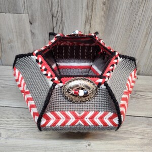May include: Handmade basket with a geometric design. The basket features a gray and black houndstooth pattern, accented with red, white, and black stitching. A silver-colored decorative clasp is on the front.