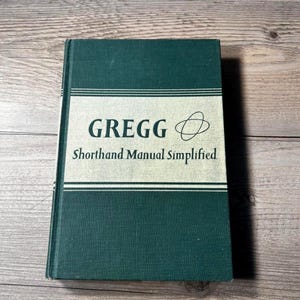 May include: A hardcover book titled "GREGG Shorthand Manual Simplified" in a dark teal color. The title is in black font on a white rectangular label with two horizontal lines. The book's cover is a solid dark teal color.