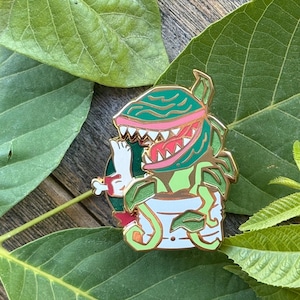May include: A gold-rimmed enamel pin featuring a green carnivorous plant with a pink interior and sharp white teeth, emerging from a white pot. The plant is holding a white bone in its mouth, with green leaves extending from the base of the pot.