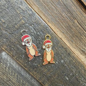 May include: Two metal charms featuring cartoon chipmunks wearing red and white Santa hats. The left charm has a silver-toned metal border, while the right charm has a gold-toned metal border. Both characters have tan bellies, brown fur, and white beards.