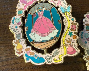 May include: A gold-toned metal enamel pin featuring a central oval frame displaying a pink dress on a hanger. The border is decorated with sewing-themed illustrations including mice, spools of thread, buttons, a measuring tape, and scissors.
