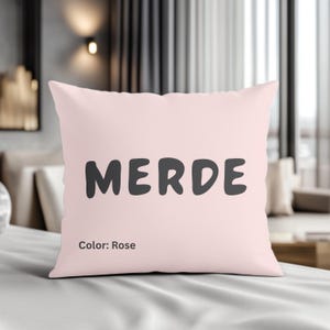 Può includere: Un cuscino quadrato color rosa con la parola "MERDE" stampata in grigio scuro. Sotto la parola c'è il testo "Color: Rose". Il cuscino è su una superficie bianca.