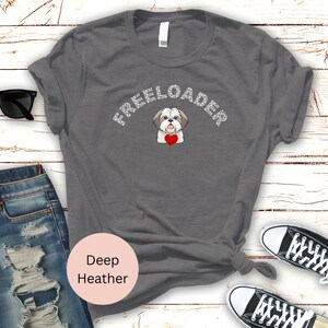 May include: Deep heather gray t-shirt with the word "FREELOADER" in white text above a cartoon Shih Tzu dog holding a red heart. The shirt is styled with jeans and sneakers.