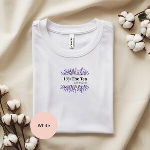 May include: White t-shirt with a crew neck. The shirt features the text "I Am The Tea" in black, surrounded by a lavender floral design. The text "-Violet Bridgerton" is below the main text. A pink circle with the word "White" is in the lower left.