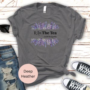 May include: Deep heather gray t-shirt with the text "I Am The Tea" and "...Violet Bridgerton" in black. The text is above and below a floral design of purple lavender. The shirt is next to a pair of sunglasses, jeans, and sneakers.