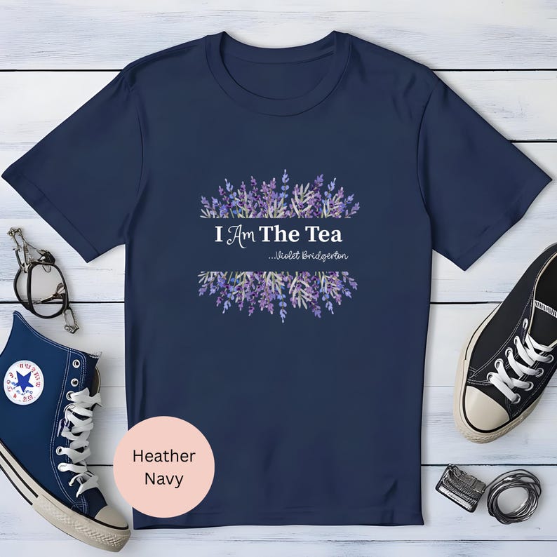May include: Heather navy t-shirt with the text "I Am The Tea" and "...Violet Bridgerton" in white. The text is framed by a watercolor design of purple lavender. A pair of navy blue sneakers and glasses are visible.
