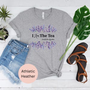 May include: Athletic heather gray t-shirt with the text "I Am The Tea" and a lavender floral design. The shirt is paired with denim shorts and brown sandals. The shirt is made of a soft, comfortable material.