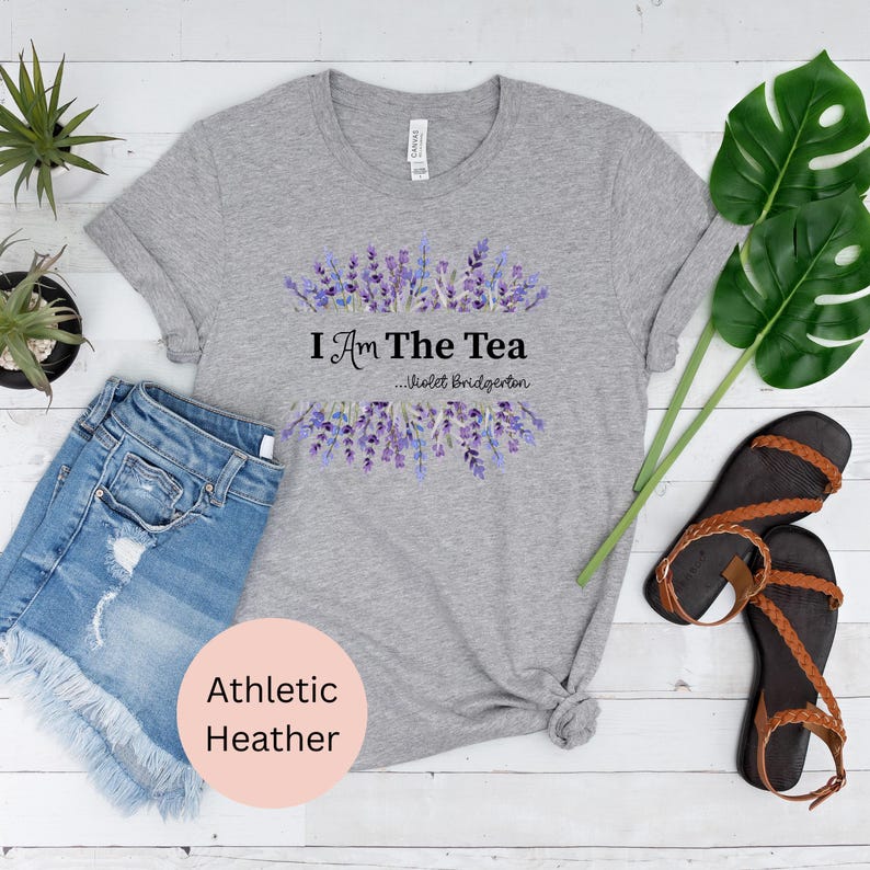 May include: Athletic heather gray t-shirt with the text "I Am The Tea" and a lavender floral design. The shirt is paired with denim shorts and brown sandals. The shirt is made of a soft, comfortable material.
