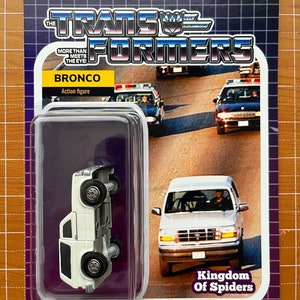 May include: A white toy car with the words "Kingdom of Spiders" printed on the bottom of the card. The car is a Ford Bronco and is part of a Transformers toy line. The card has a warning label that says "choking hazard" and "adult collectible - not a toy".