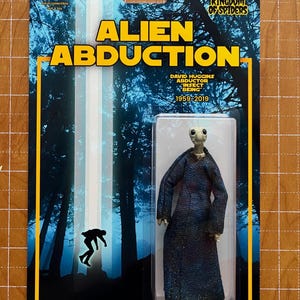 Puede incluir: Figura de acción coleccionable titulada "Alien Abduction". La figura es de un alienígena con una larga túnica azul, empaquetada en una tarjeta con imágenes de bosque y el texto "Alien Abduction". La tarjeta también incluye el texto "Kingdom of Spiders".