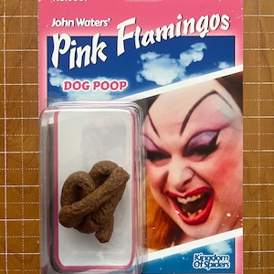Puede incluir: Un artículo de novedad empaquetado en un blíster rosa y azul. El paquete presenta el texto "John Waters' Pink Flamingos DOG POOP" y una imagen de una persona con maquillaje dramático. Se ve una caca de perro marrón esculpida.