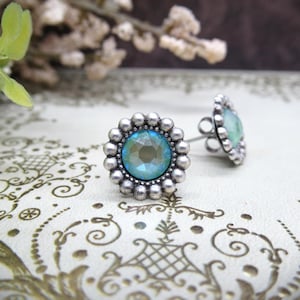 May include: A pair of silver-toned stud earrings. Each earring features a central, faceted, light blue gemstone surrounded by a beaded border. The earrings are set against a decorative background, with one earring in focus and the other slightly blurred.