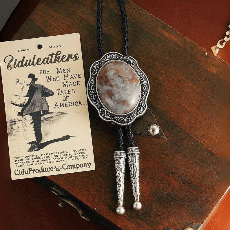 May include: A bolo tie with a large, oval, marbled stone pendant set in a decorative silver-tone frame. The black braided cord has silver-tone tips. An antique advertisement for Biduleathers is in the background.